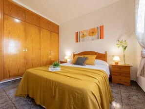 Une chambre avec une armoire en bois, un lit recouvert d’une couverture jaune, une table de chevet avec une lampe et un tableau accroché au mur.