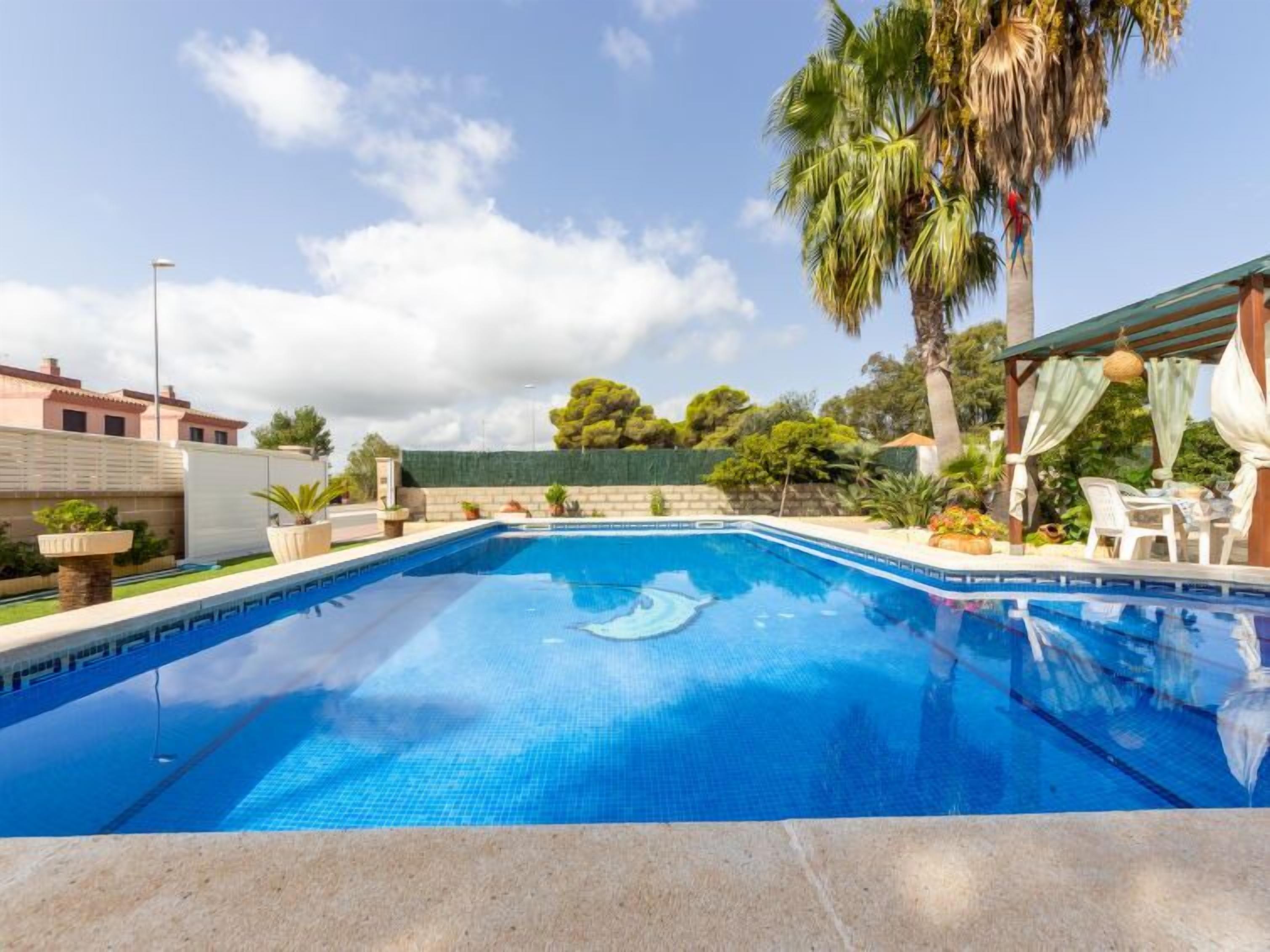 Una piscina rectangular con agua azul cristalina, rodeada por una terraza de piedra. Hay plantas en macetas y una pérgola con cortinas blancas en un lado.
