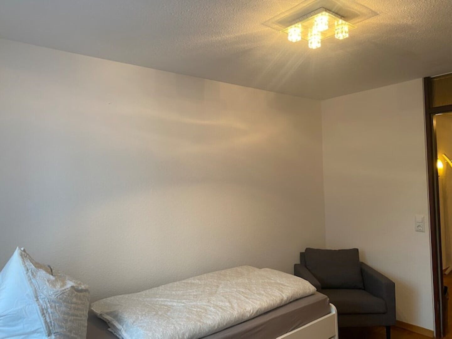 A small, sparsely furnished room with a single bed, a chair, and a door leading to another room.