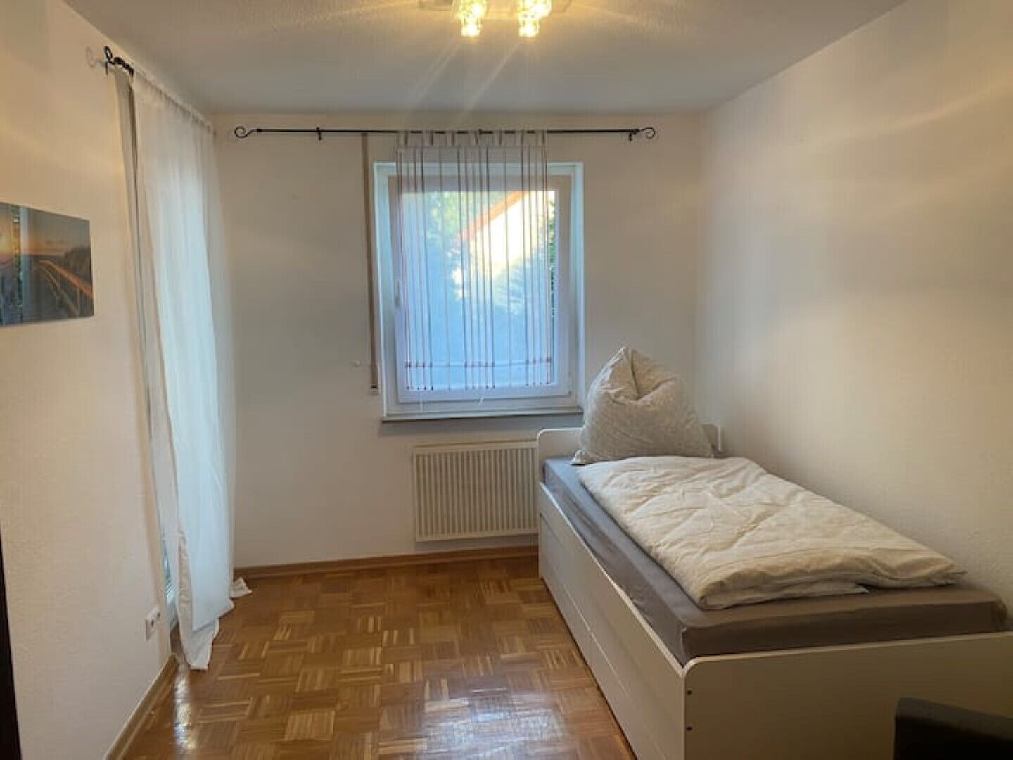 A small, single-bed room with a wooden floor, a window with blinds, a radiator, and a ceiling light fixture.