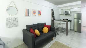 Una sala de estar moderna con un sofá negro, un espejo redondo y varias obras de arte en la pared. Se observa una zona de cocina con microondas y refrigerador.