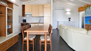 A modern kitchen with wooden cabinets, a dining table with chairs, and a white sofa in a bright room with a tiled floor. - Arbatax Sa Omu Bianca (Tortoli)