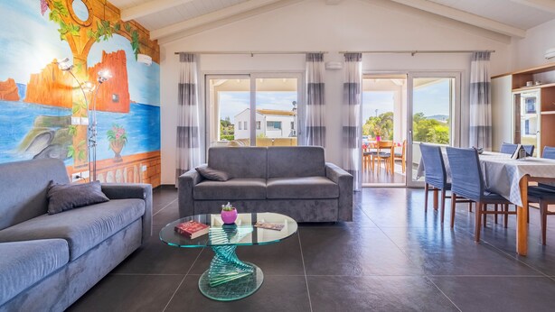 A spacious living area with a grey sofa, a glass coffee table, and a mural on the wall. - Arbatax Sa Omu Bianca (Tortoli)