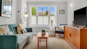 A modern living room with a sofa, a coffee table, a TV, and a large window with a view of greenery. - Charming cottage in the Heart of Whitstable - Pass the Keys (Whitstable)