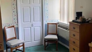 Une chambre à coucher comprenant un lit, deux chaises, une commode, un radiateur et une porte ornée d’un motif floral.