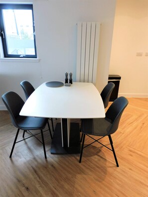 Une salle de conférence avec une table blanche, des chaises noires, un petit ensemble de salière et de poivrière, et une fenêtre avec des stores.