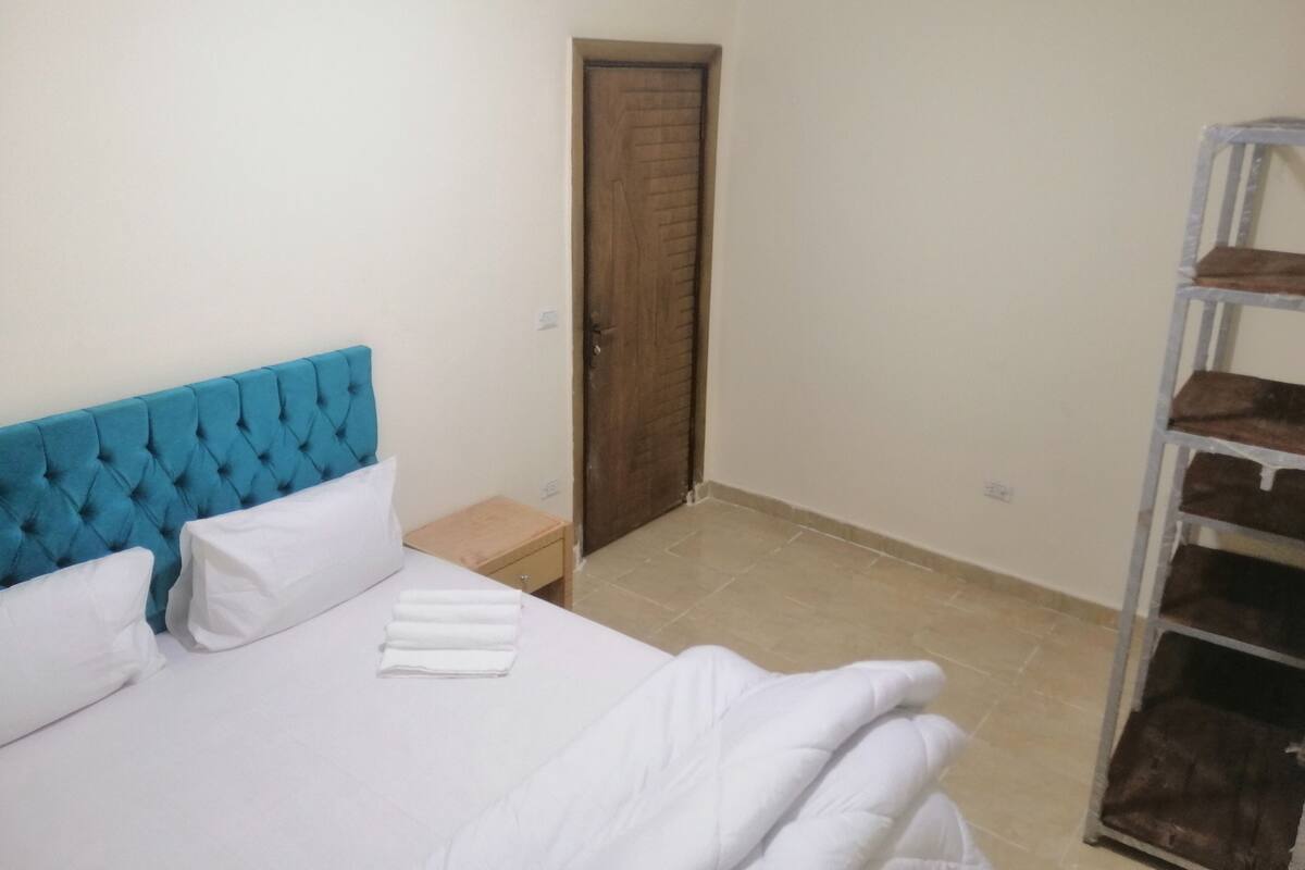 A bedroom with a blue tufted headboard, white bedding, a wooden door, and a metal shelf unit.