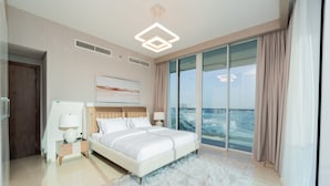 A modern hotel room with a large bed, a nightstand, a lamp, a painting, and a view of the city through large windows. - GLOBALSTAY. New Apartments in Business Bay (Dubai)