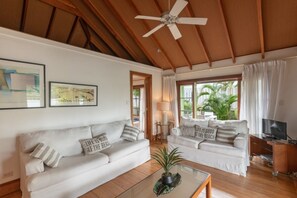 Un salon avec deux canapés blancs, un ventilateur de plafond, une table basse ornée d’une plante et un meuble pour la télévision.