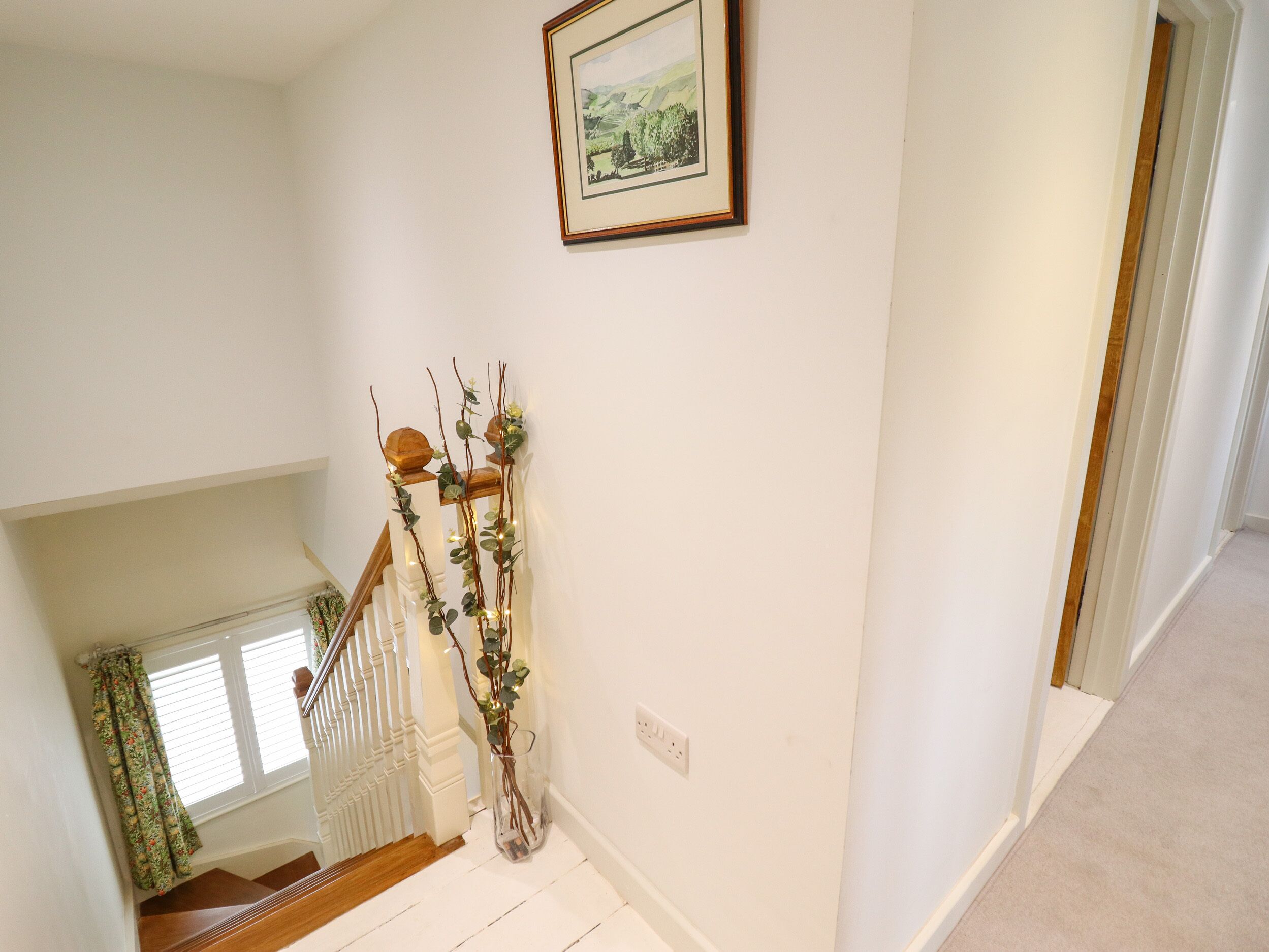 A staircase with a glass vase of greenery, a framed landscape painting, and a window with patterned curtains.