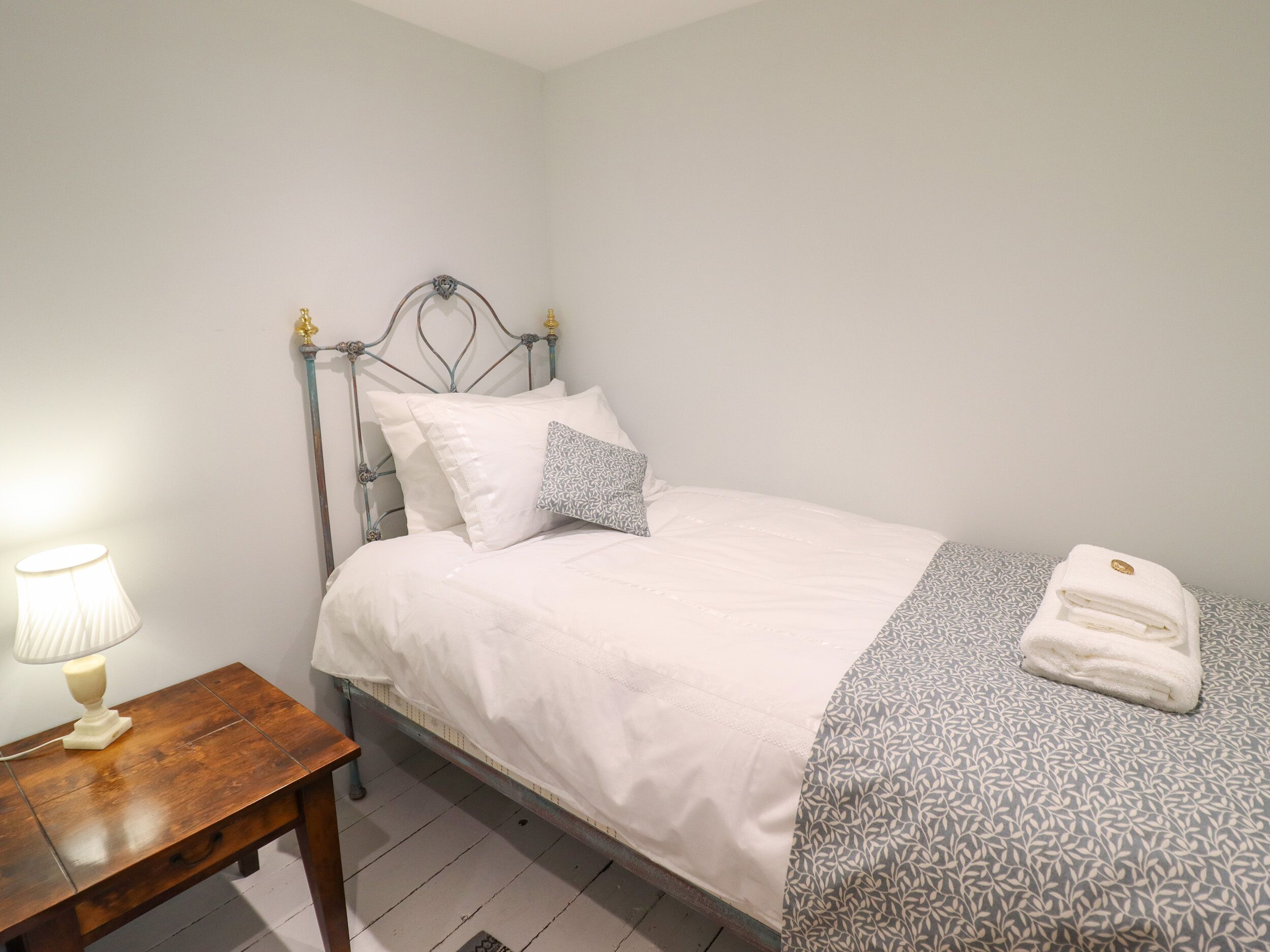 A neatly made bed with white bedding and a patterned throw, a bedside table with a lamp, and a wooden nightstand.