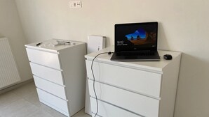 A room with a white desk and a laptop on it, a white radiator, and a white cabinet with drawers.