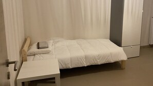 A single bed with a wooden headboard, a bedside table, and a wardrobe in a room with plain walls and a tiled floor.