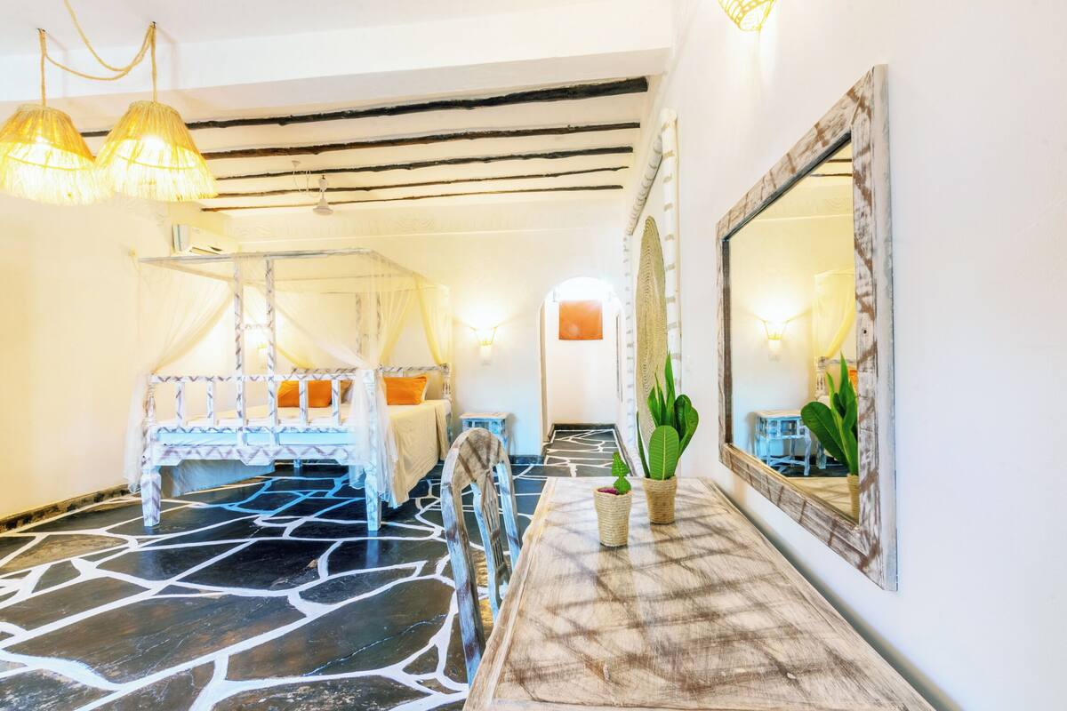 A room with a four-poster bed, a wooden dining table with two potted plants, and a large mirror on the wall.