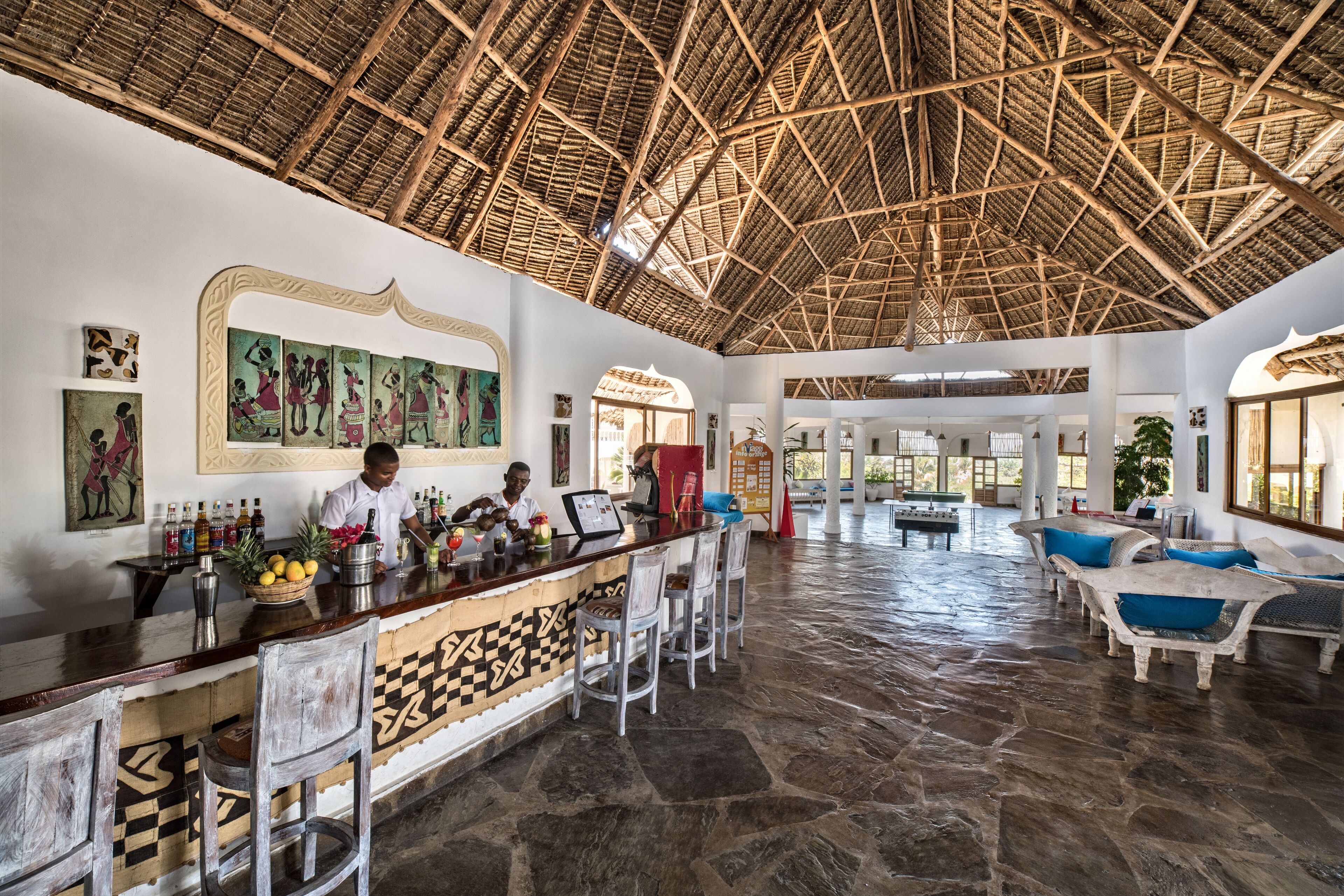 Eine großzügige Lobby mit einer hohen Strohdecke, einer Bar mit Hockern und einer Sitzecke mit blauen Kissen.