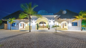 Un resort con entrada de techo de paja, palmeras y un camino pavimentado.