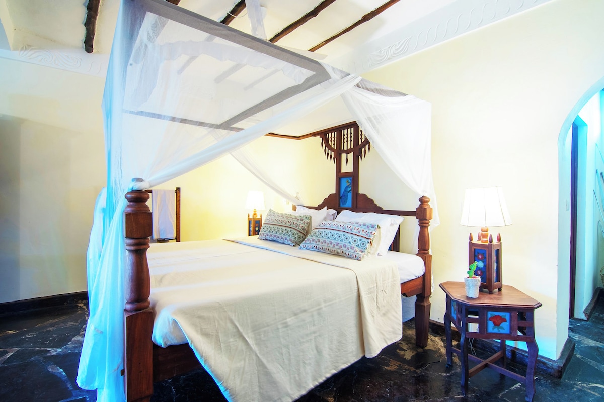 A four-poster canopy bed with a wooden frame and white bedding. A small table with a lamp and decorative items is beside the bed.