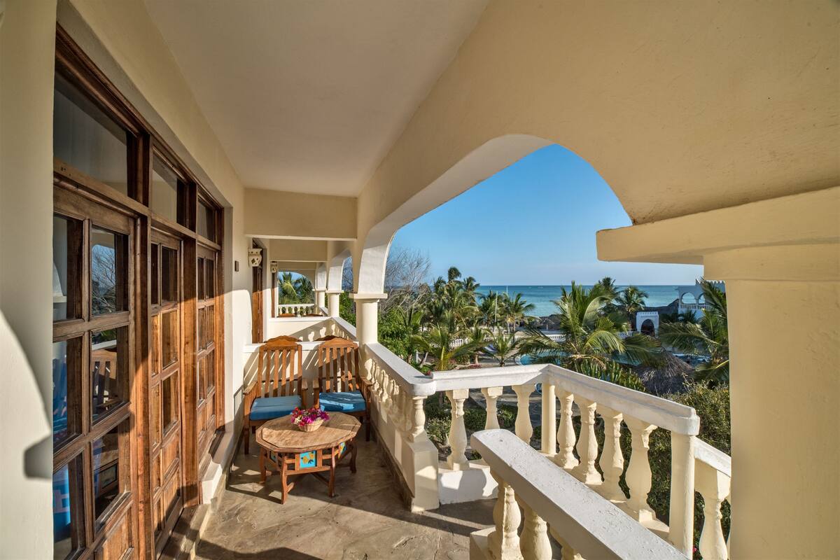 A balcony with a view of the ocean, wooden furniture, and a table with a vase.