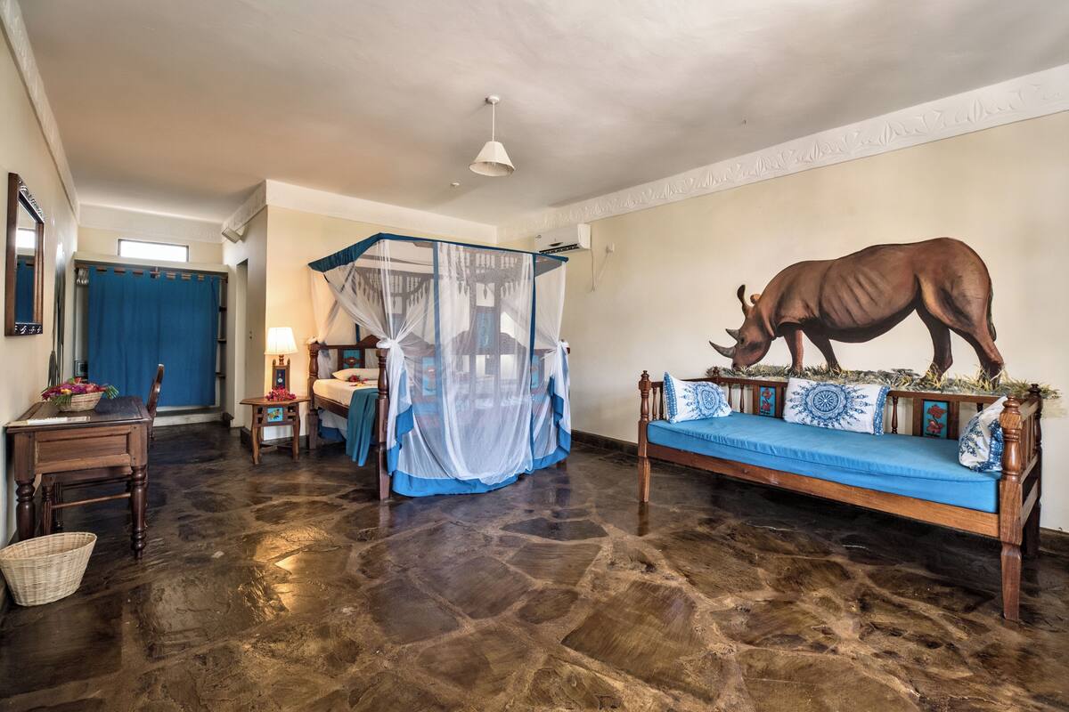 A room with a stone floor, a bed with a blue canopy, a wooden sofa with decorative pillows, a wall-mounted rhinoceros sculpture, and a small table with a lamp.