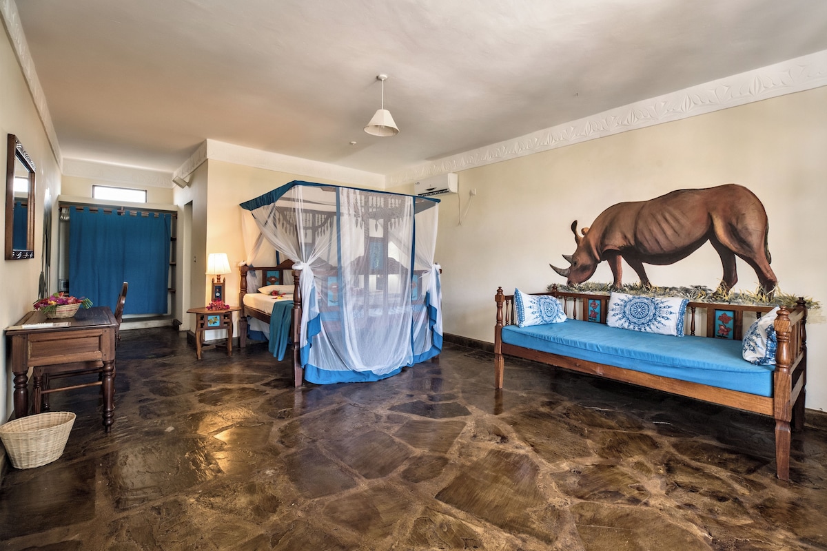A room with a stone floor, a bed with a blue canopy, a wooden sofa with decorative pillows, a wall-mounted rhinoceros sculpture, and a small table with a lamp.