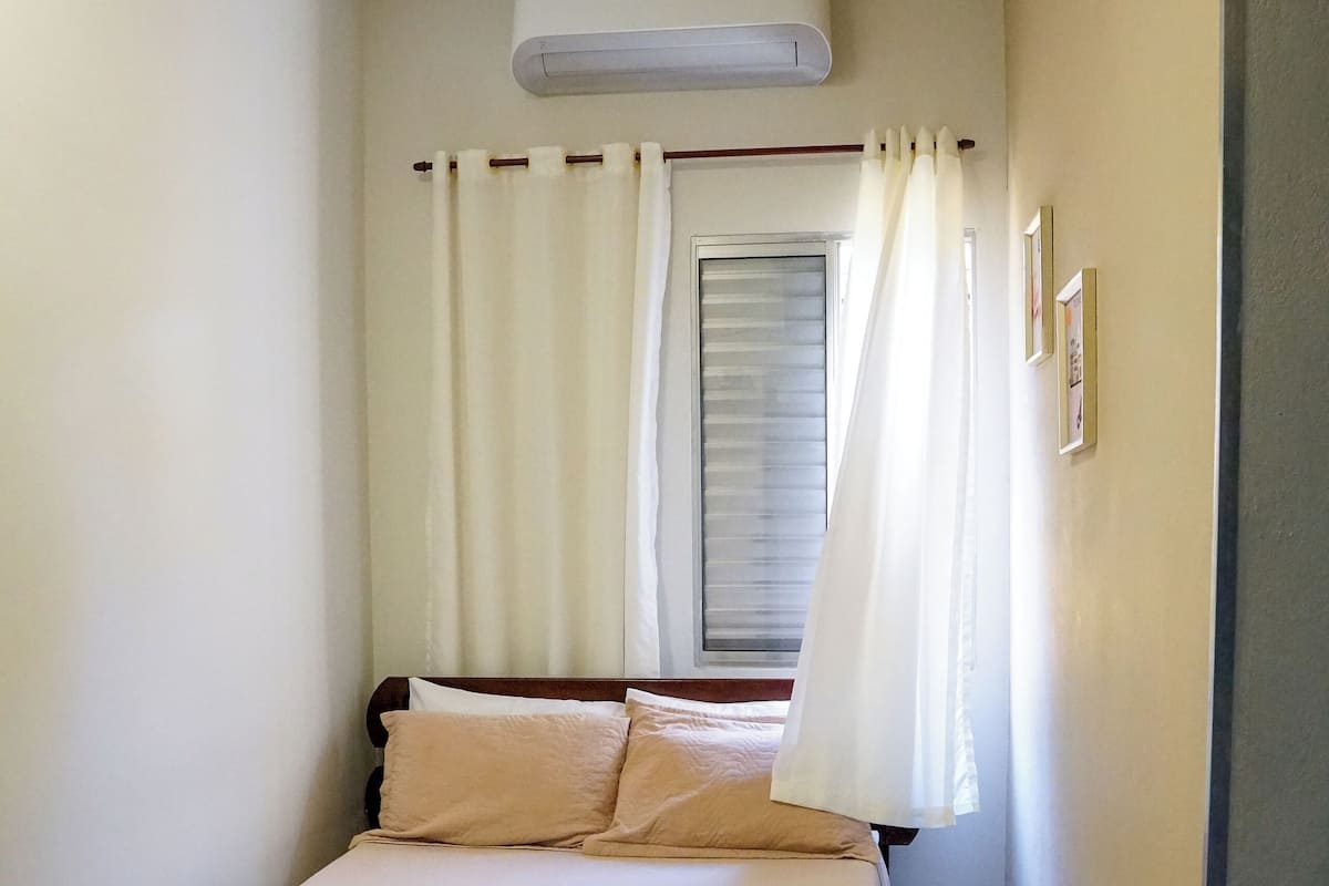 A small, neatly arranged bedroom with a single bed, a window with curtains, and an air conditioning unit above it.