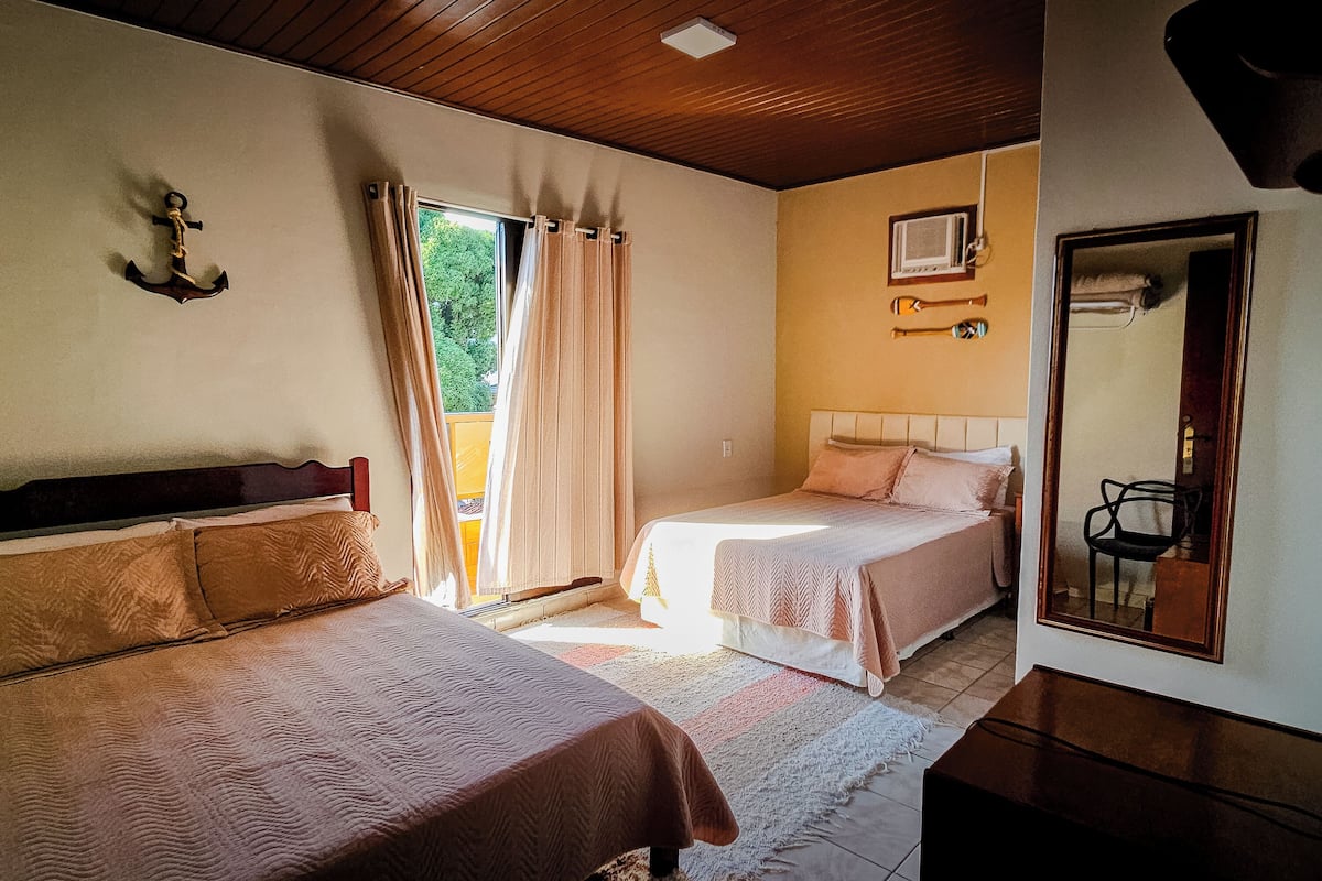 A bedroom with two single beds, a wooden ceiling, a window with curtains, a small bedside table, and a mirror on the wall.