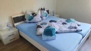 Siististi pedattu sänky, jossa on sininen päiväpeite ja koristeellisia tyynyjä, yöpöytä lampun kanssa ja puulattia.