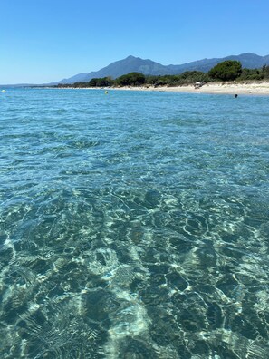 Klares, türkisblaues Wasser mit erkennbaren Details unter der Oberfläche, ein Sandstrand und eine Bergkette im Hintergrund.