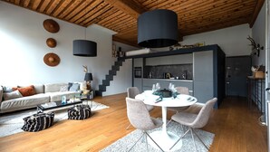 A modern living room with a wooden ceiling, a dining area with a round table and chairs, and a staircase leading to a kitchen area. - LE LOFT ETHNIC - Grand Studio avec magnifique vue (Lyon)