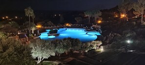 Une grande piscine illuminée, entourée de chaises longues et de parasols, aménagée dans un espace paysager agrémenté d’arbres et d’un belvédère.