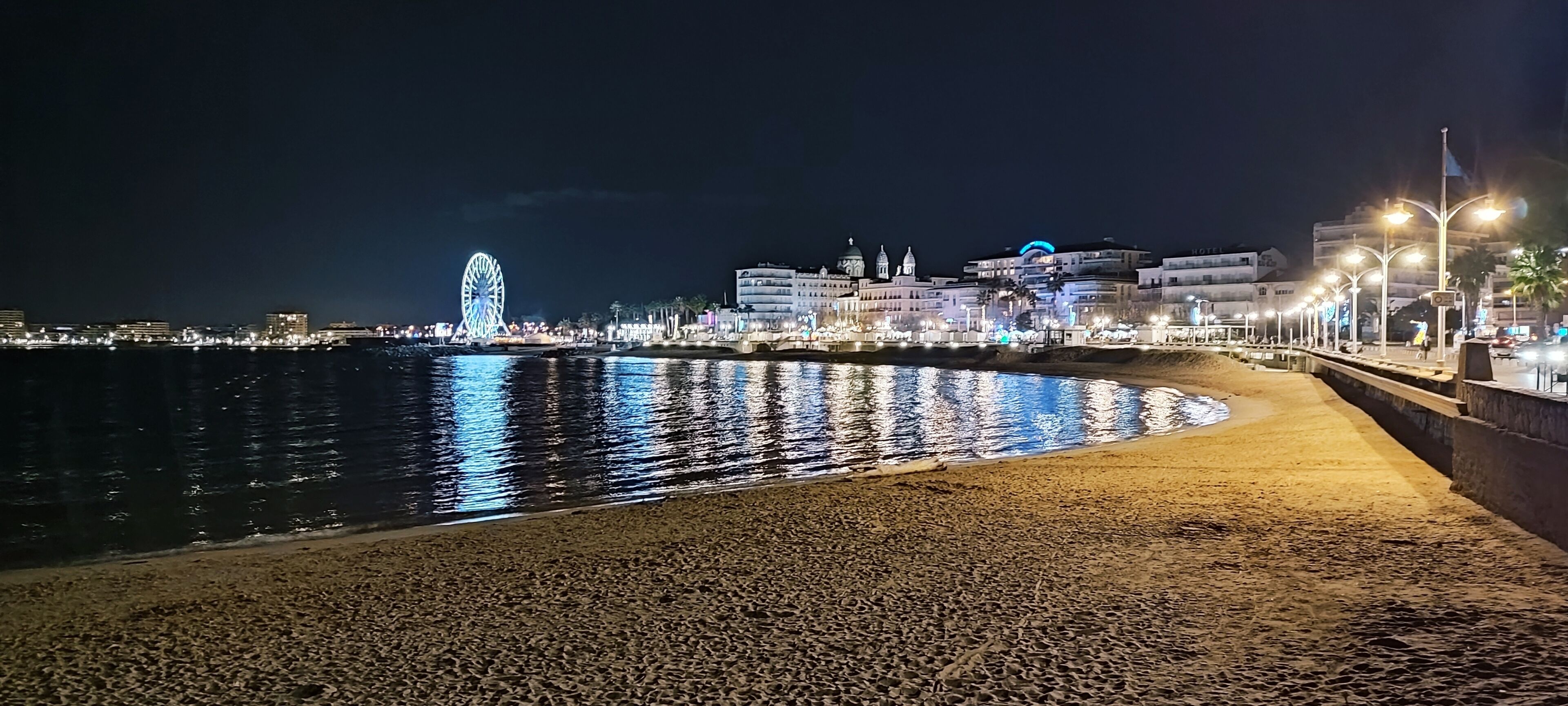 A coastal cityscape at night with a prominent Ferris wheel, illuminated buildings, and a sandy beach with a promenade.