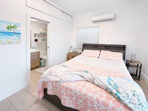 A bedroom with a bed, a nightstand, a bedside table, a window with blinds, an air conditioning unit, and a door leading to a bathroom. - Prívate suite in Calusa Campgrounds (Key Largo)
