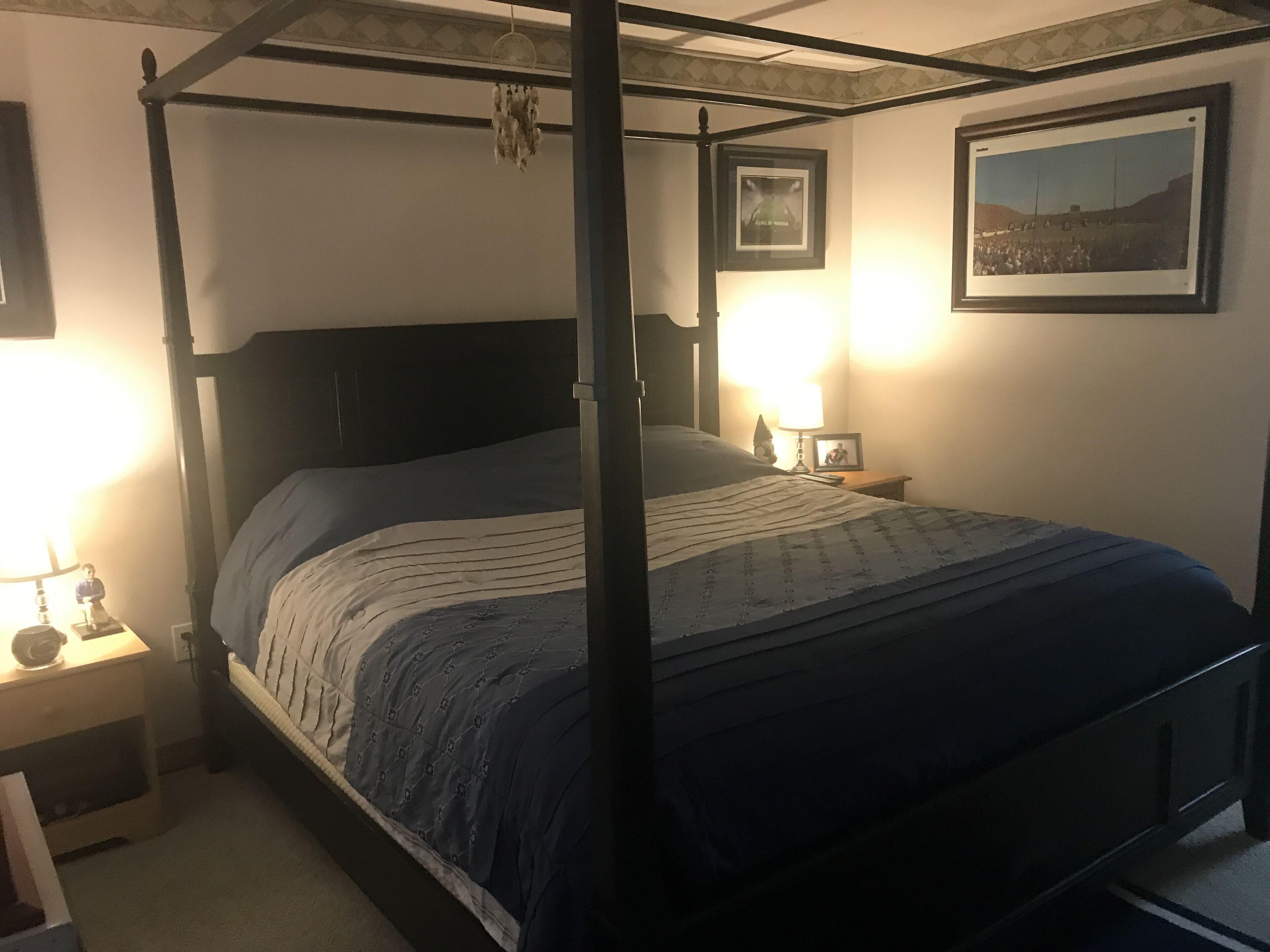Una cama con dosel, un cabecero oscuro y una colcha acolchada. Hay dos mesitas de noche con lámparas y cuadros enmarcados. La habitación tiene un diseño sencillo y funcional, con una pared lisa y un cuadro sobre la cama.