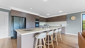 A modern kitchen with a central island, bar stools, a refrigerator, oven, microwave, and a clock on the wall. - Blue Haven, North Narooma (North Narooma)