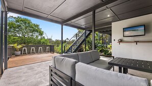 A modern outdoor living area with a wooden deck, grey sofa, and a table. - Blue Haven, North Narooma (North Narooma)