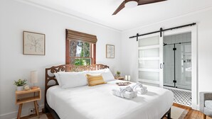 Quarto bem organizado com uma cama grande, uma mesa de cabeceira com abajur, janela com persianas e uma porta de vidro corrediça que dá para um espaço com chuveiro.