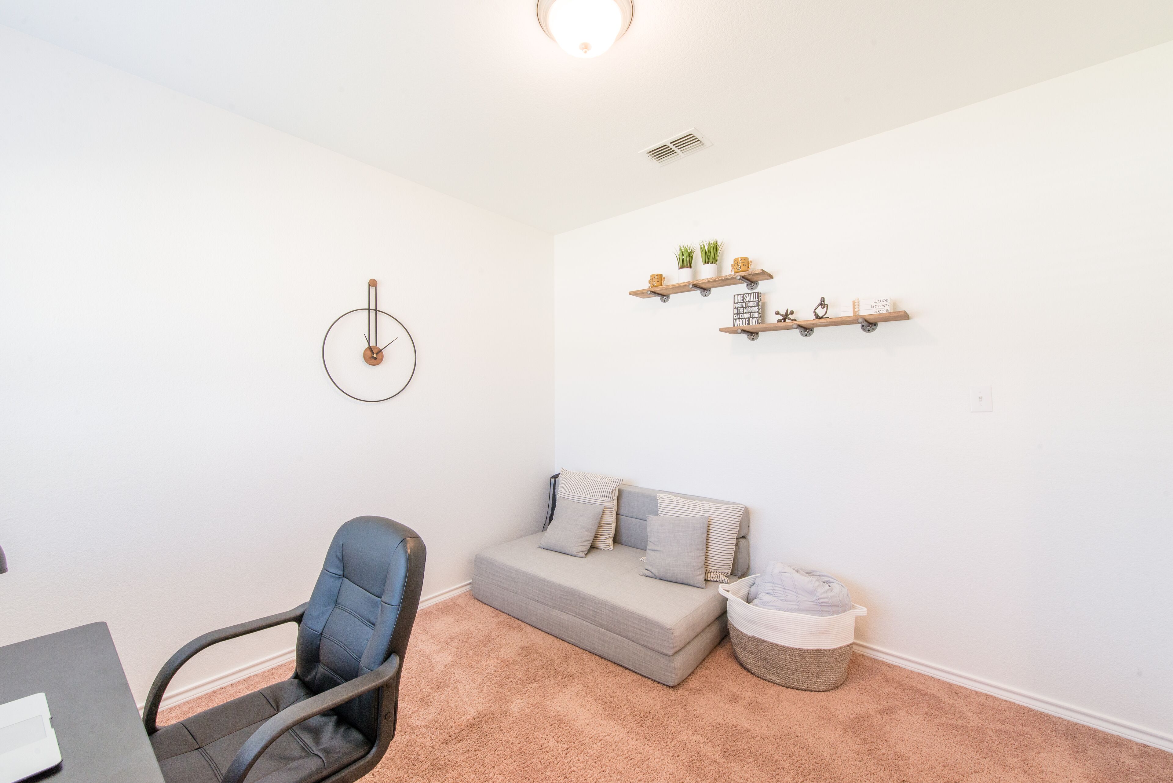 A room with a grey sofa, a blue office chair, a small table, and a wall-mounted shelf with decorative items.