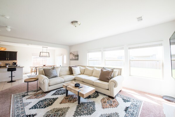 A spacious living area with a large L-shaped sofa, a coffee table, and a patterned rug. There is a kitchen area in the background with a bar stool and a dining table. - A Spacious And Fully Stocked House (Fort Worth)