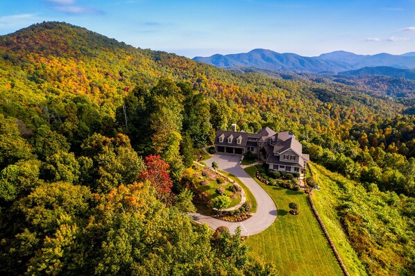 A large, modern house with a spacious driveway and landscaped garden, surrounded by a forested mountain landscape.