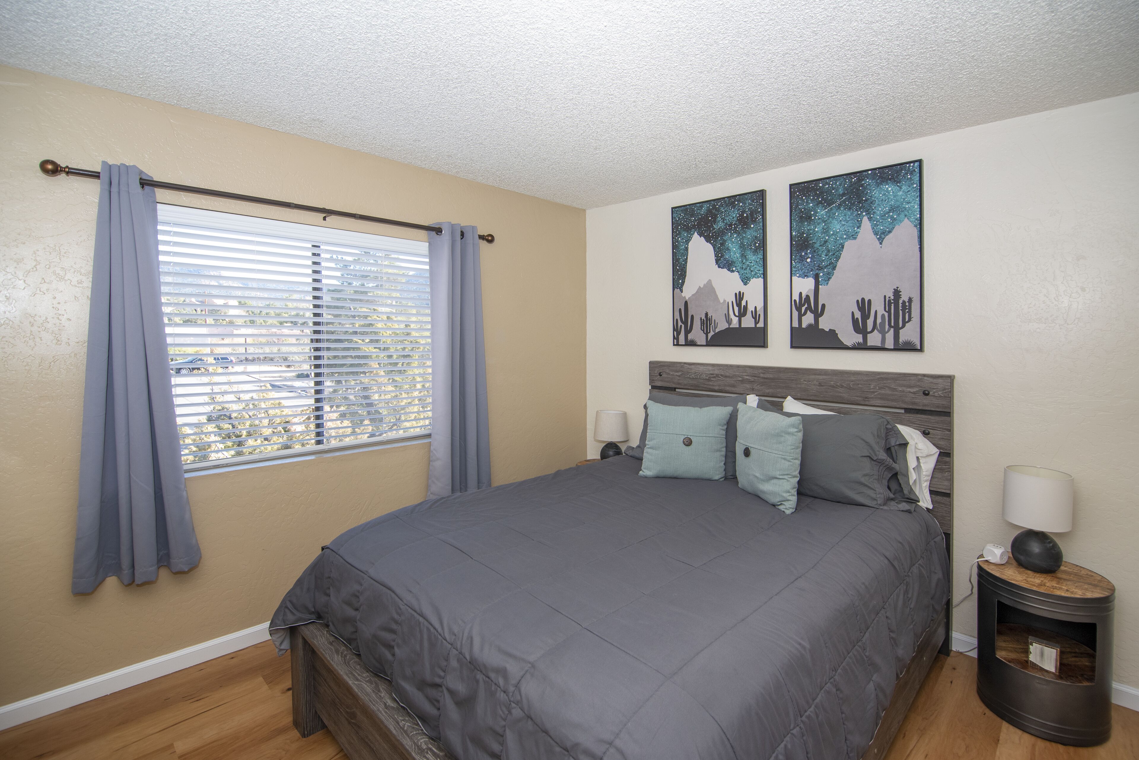 A bedroom with a large bed, a window with blinds, and two wall art pieces depicting mountains and stars.