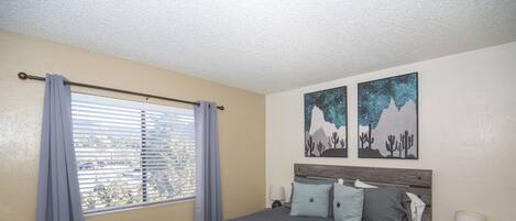 A bedroom with a large bed, a window with blinds, and two wall art pieces depicting mountains and stars.