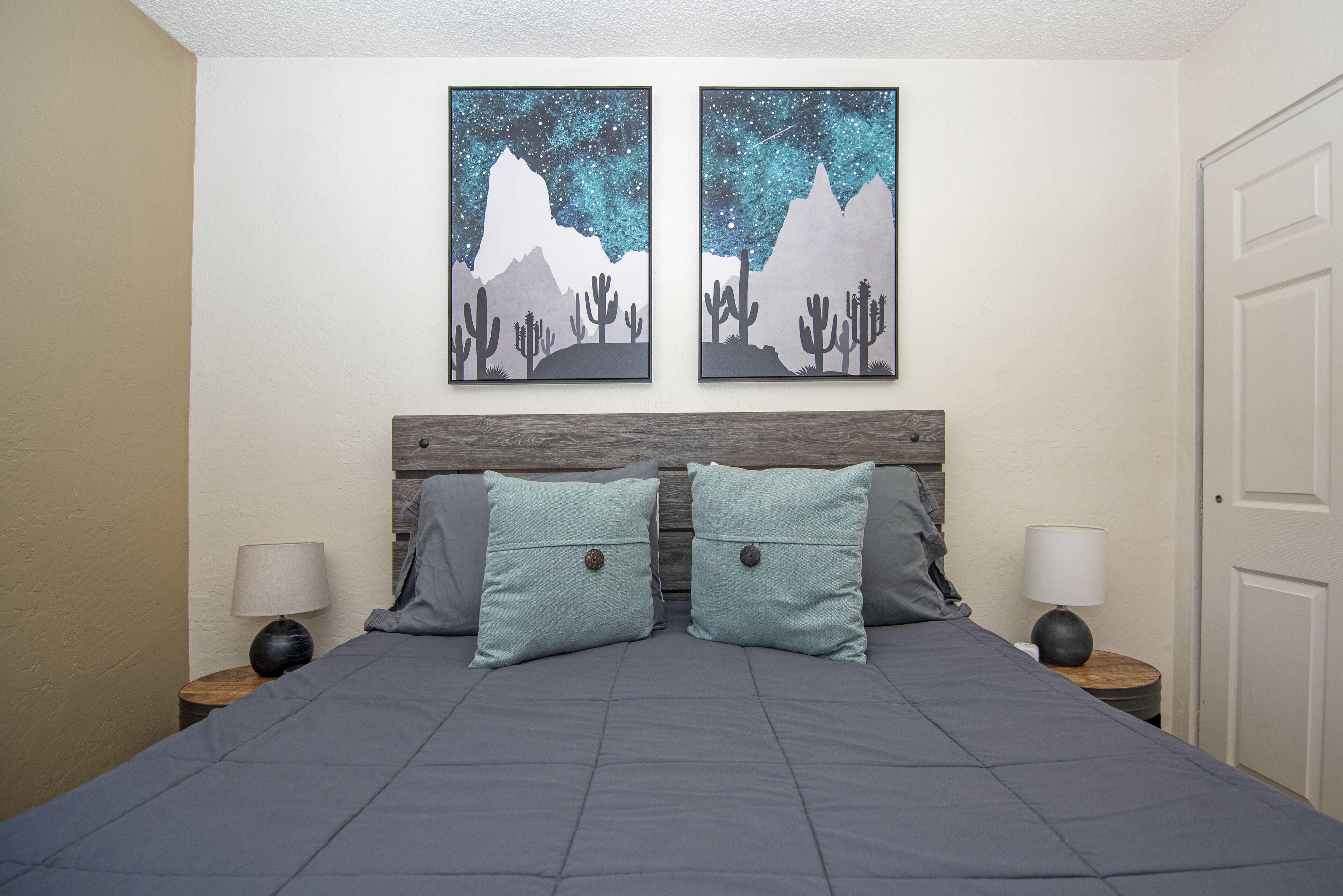 A bedroom with a bed, two pillows, two nightstands, two lamps, and two wall art pieces depicting a starry night sky and mountains.