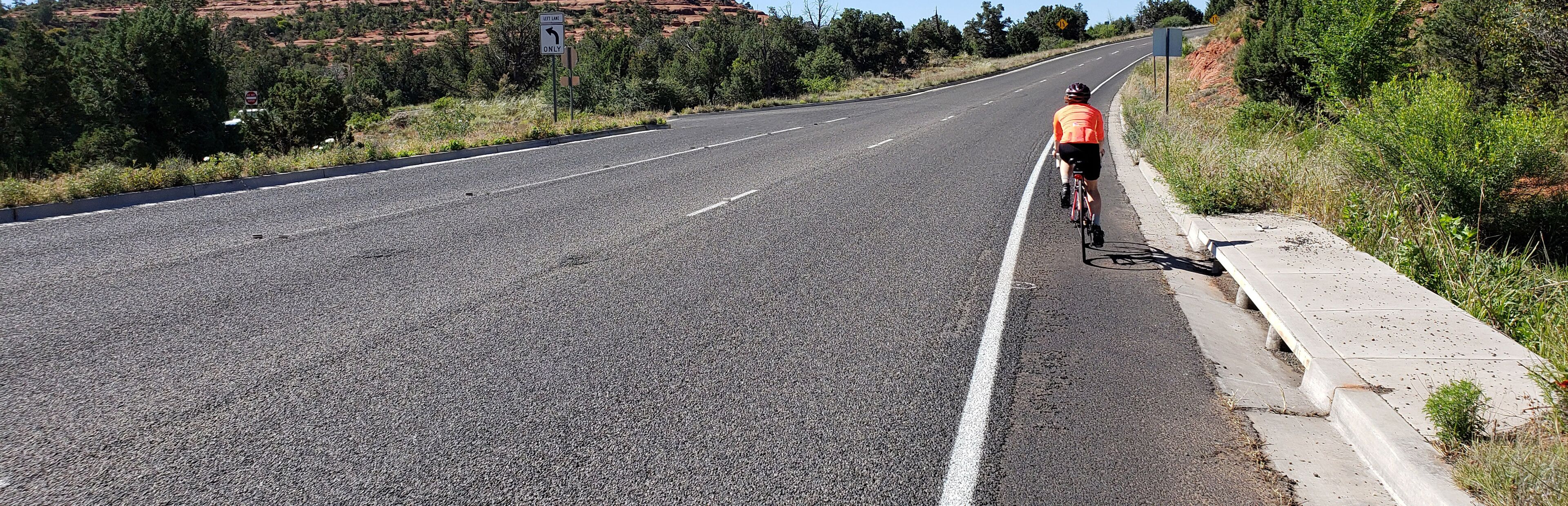 Un ciclista pedaleando por una carretera con un cartel indicador de giro a la izquierda, rodeado de follaje y colinas.