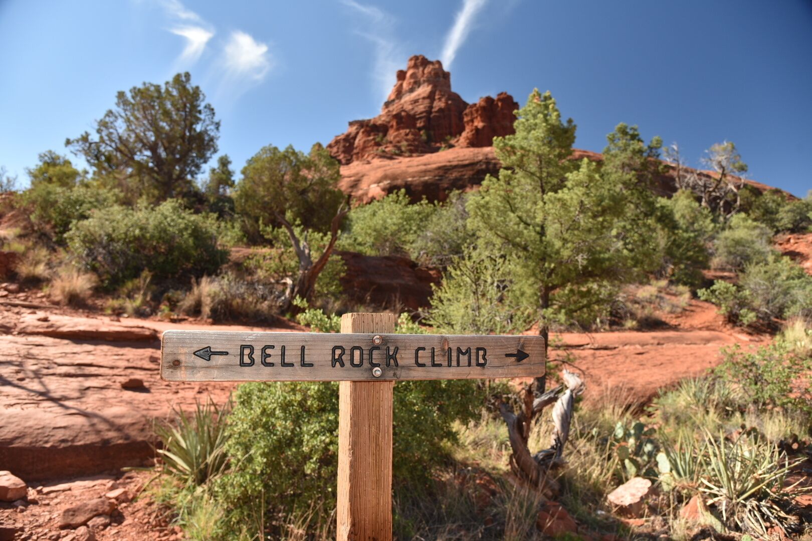 A wooden signpost with directions to 'Bell Rock Climb' in a desert landscape with red rock formations and sparse vegetation.