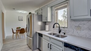 A modern kitchen with white cabinets, a stainless steel refrigerator, a dishwasher, and a window with blinds. - Beautiful 3BR Townhouse (Augusta)