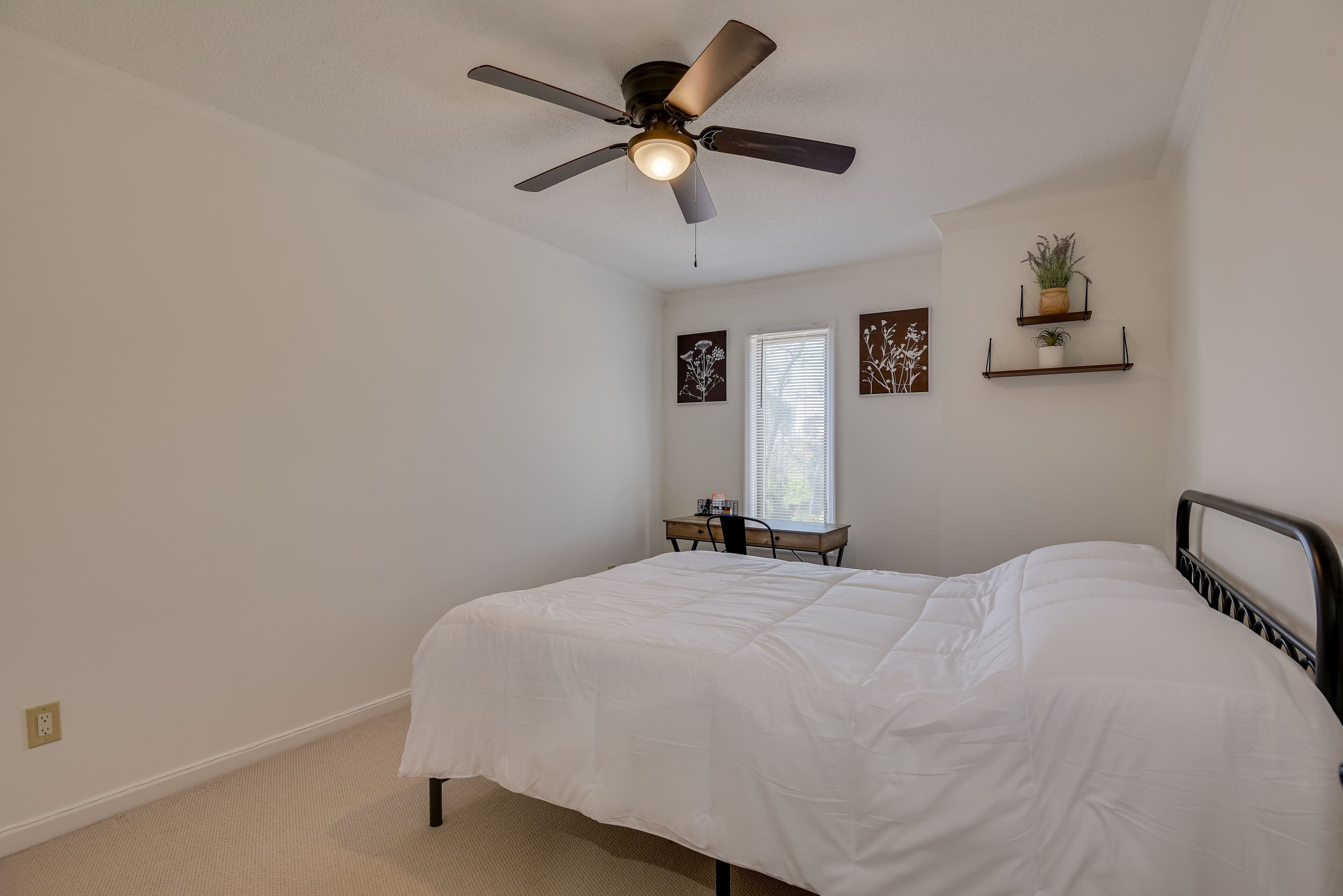A bedroom with a bed, a ceiling fan, a desk, and two wall-mounted shelves with plants.