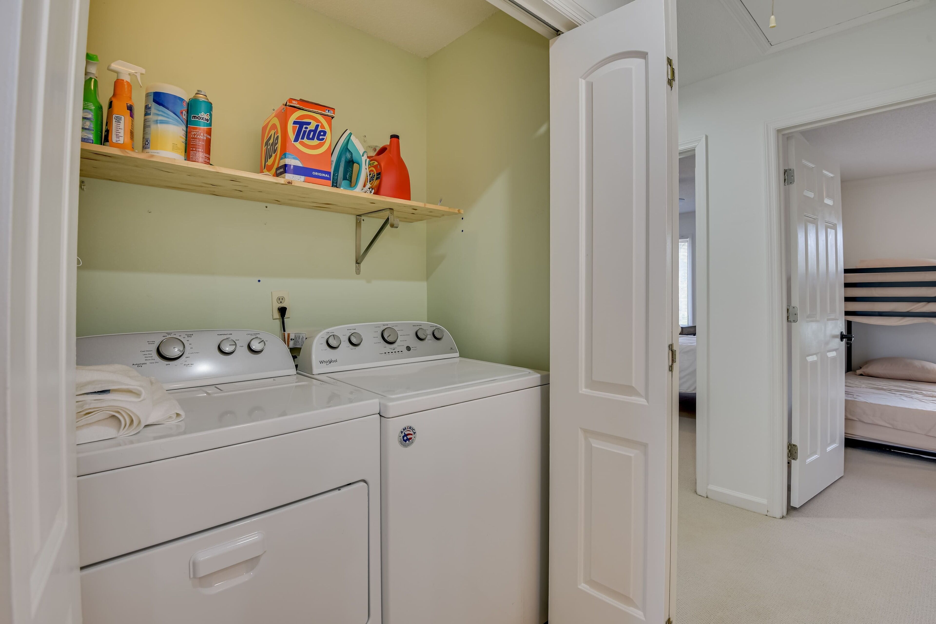 A laundry area with a washing machine and dryer, a shelf with cleaning products, and a bunk bed in the background.
