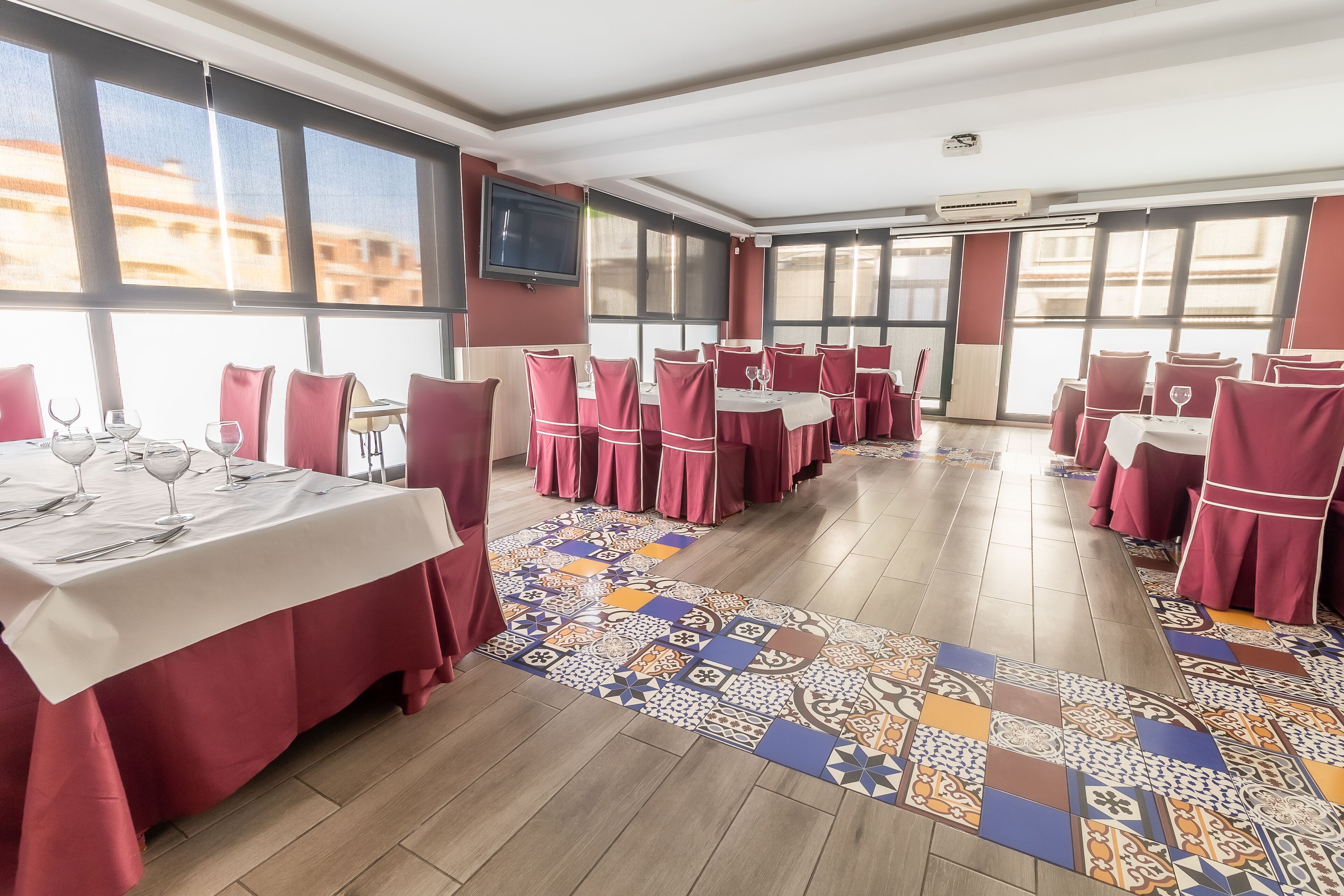 Comedor con mesas dispuestas para el servicio, ventanas amplias y alfombra estampada.