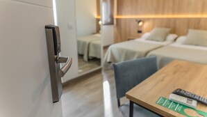 A hotel room with a modern design, featuring a wooden floor, a bed with a white headboard, a small table with a remote control, and a door with a digital lock. - Hotel Kika (Santa Marta)