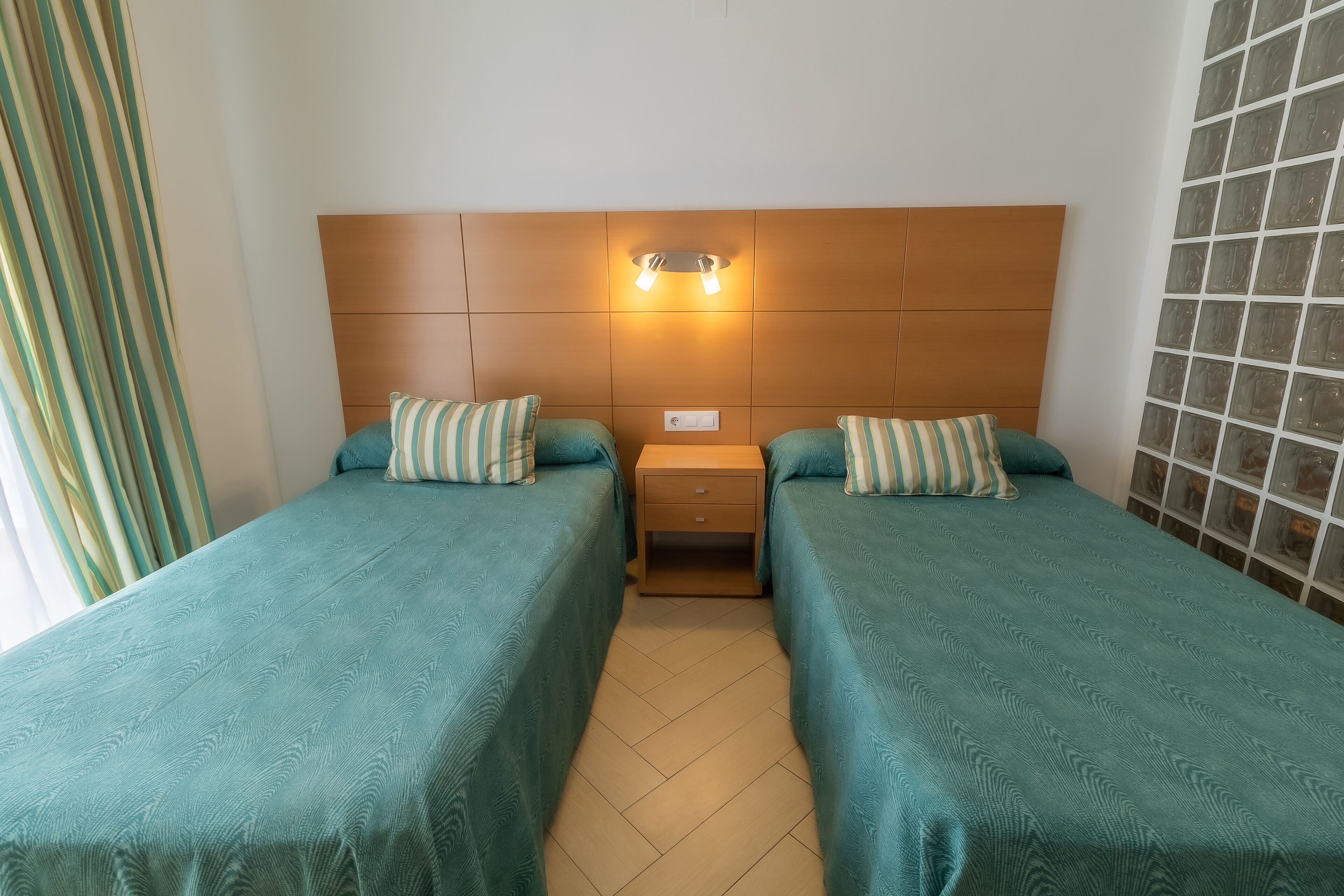 Habitación con dos camas, cabecera de madera, mesita de noche y pared con diseño de bloques de vidrio.